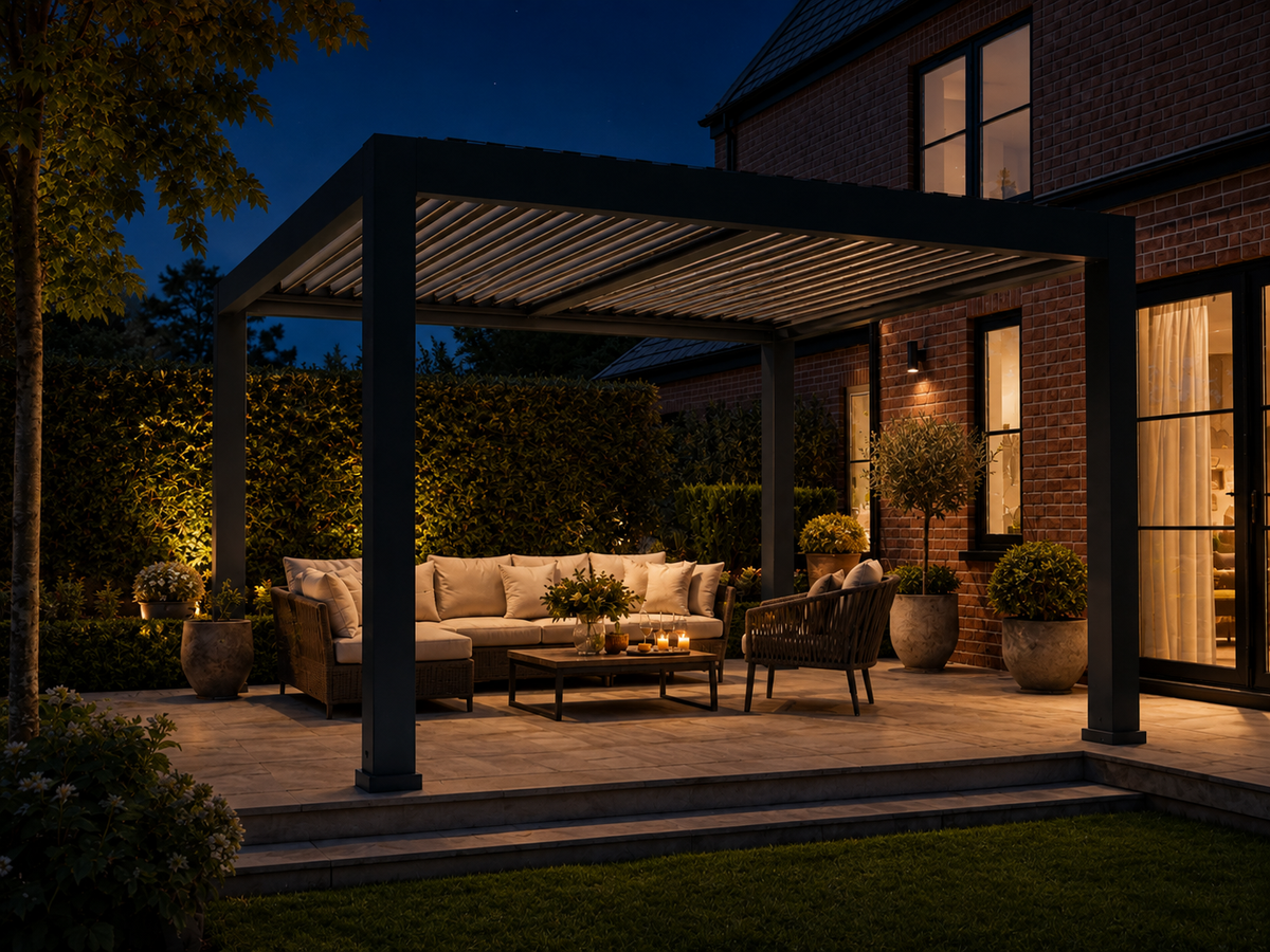 Outdoor patio at dusk with a dark pergola structure over a furnished seating area, including a beige sectional sofa, coffee table, and chairs, beside a brick house with lit windows.